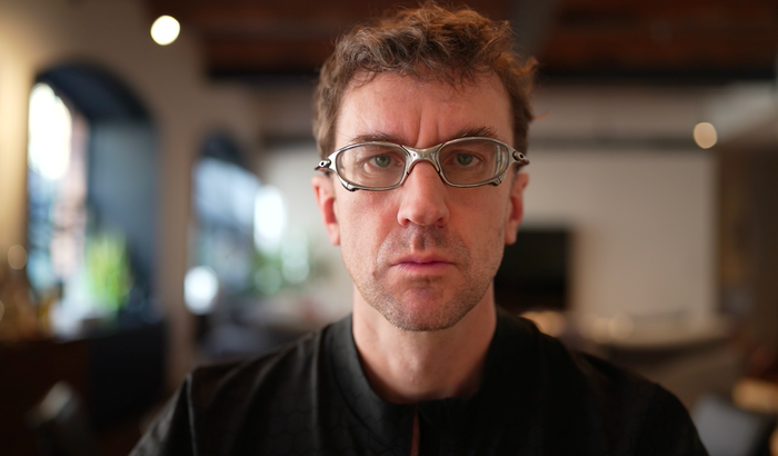 Photo of inventor Pablos Holman wearing silver glasses and a black shirt, looking straight at the camera. The background is a blurred room.